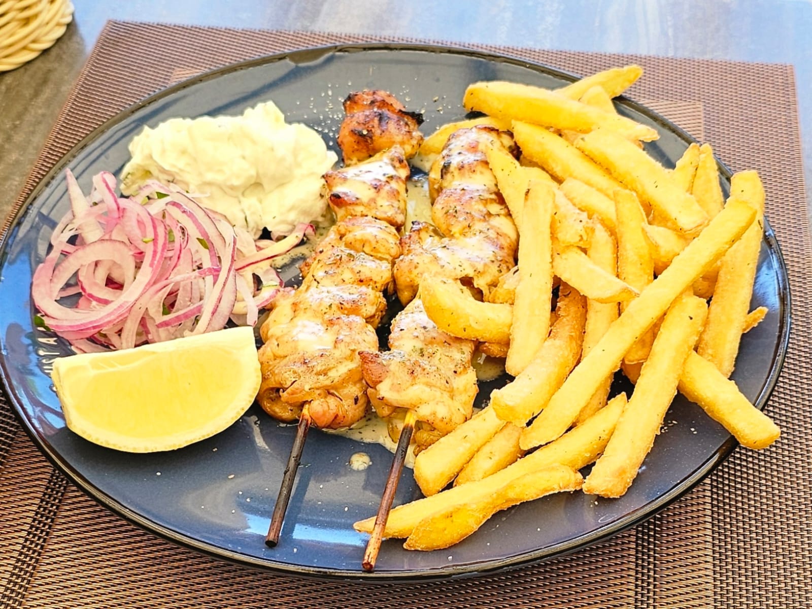 Ein dunkler Teller mit zwei Hähnchenspießen, Pommes, Zwiebeln, Zitrone und Dip
